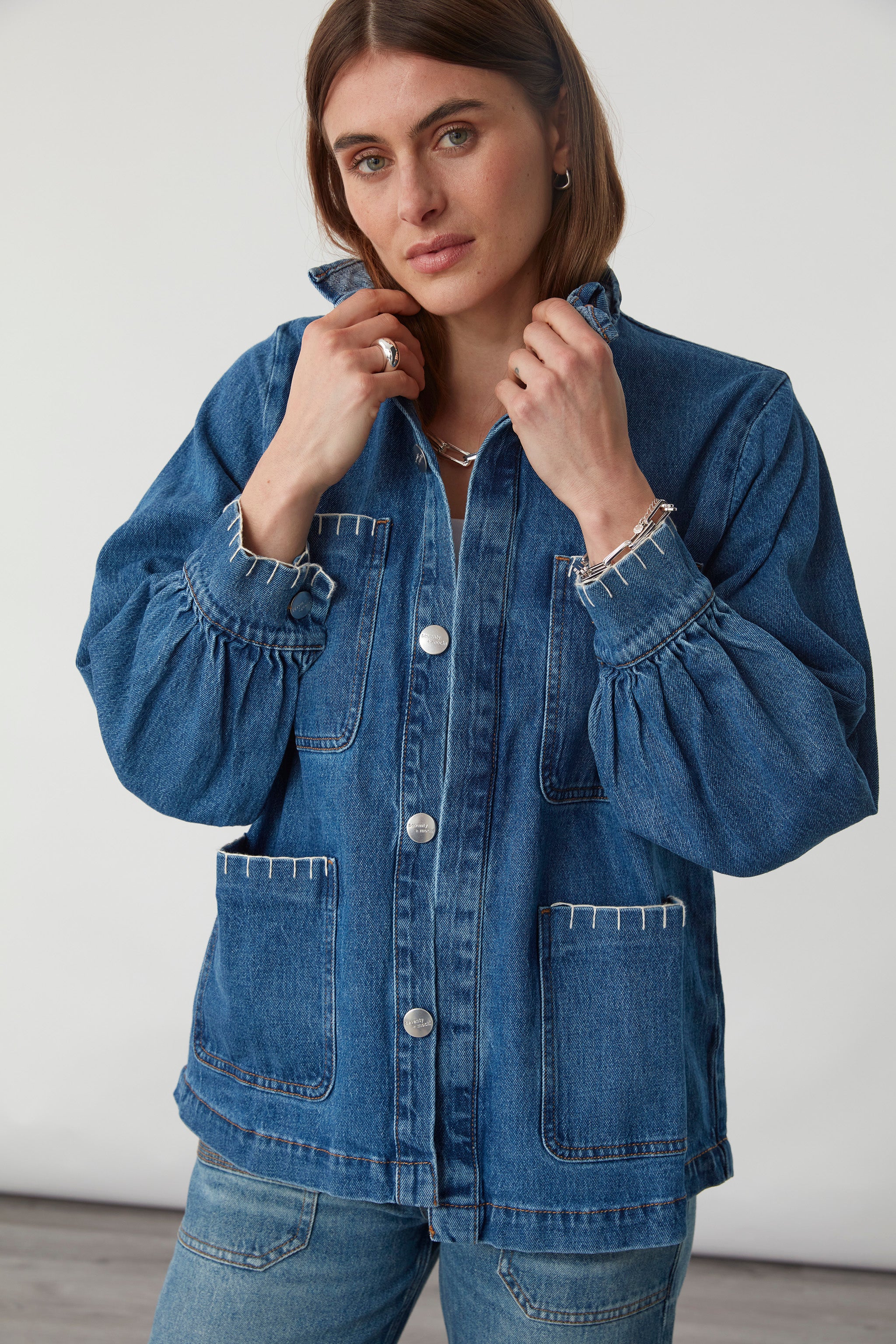 woman wearing a blue denim jacket with a frill collar and white blanket stitching on four large patch pockets and blue jeans against a plain background