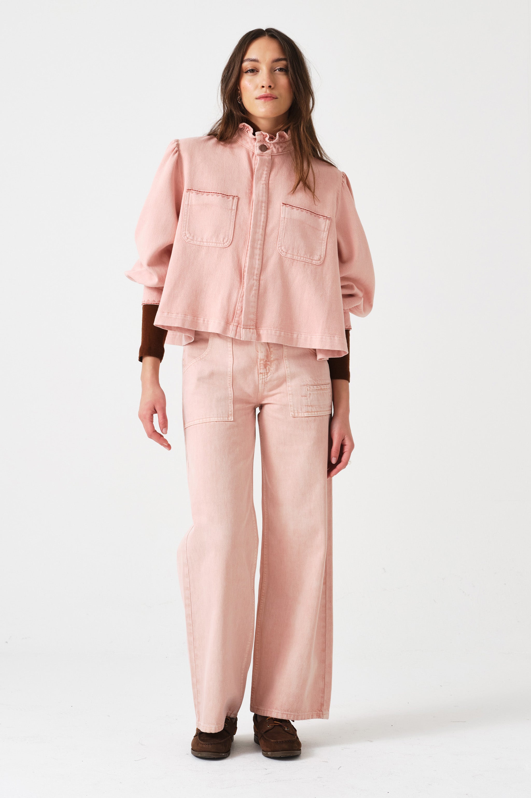 woman wearing light pink high waisted, wide, cropped jeans with big pockets, a pink shirt, and brown shoes on a white background