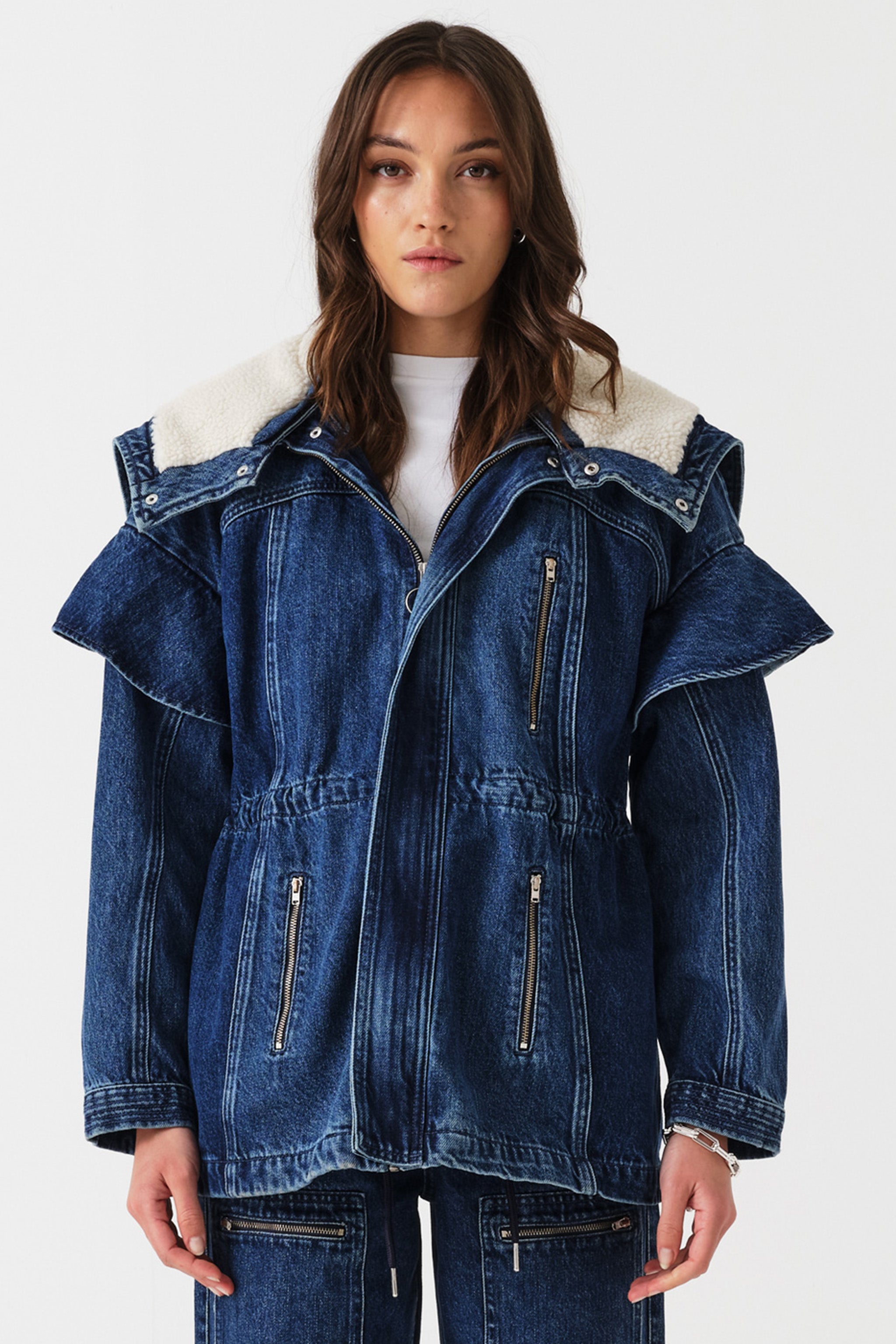 Woman wearing a dark blue denim jacket with a white sherpa lined removable hood, waist cinch, and zip pocket details on a white background