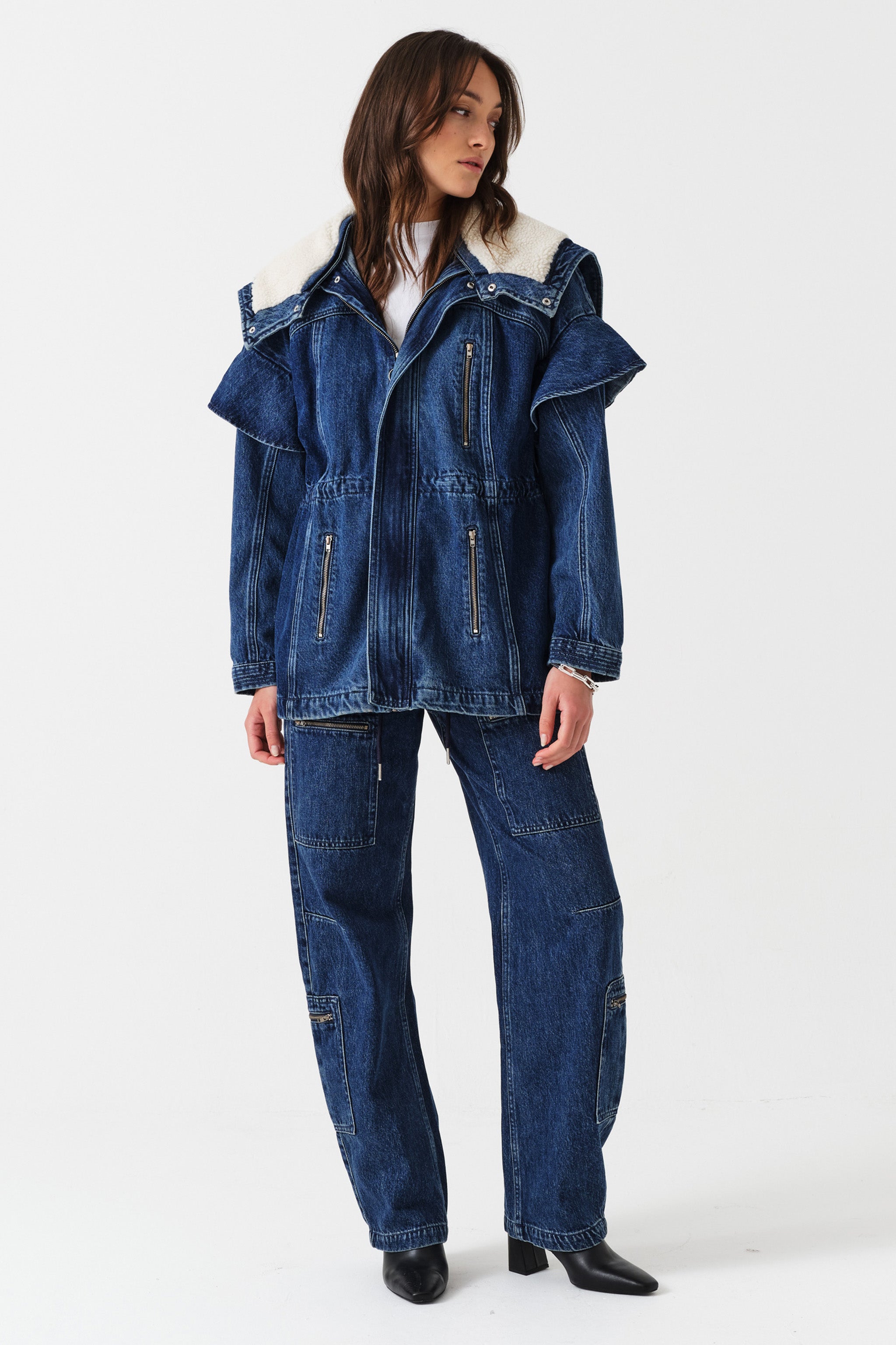 Woman wearing a dark blue denim jacket with a white sherpa lined removable hood, waist cinch, and zip pocket details with dark blue jeans with cargo zip pockets on a white background