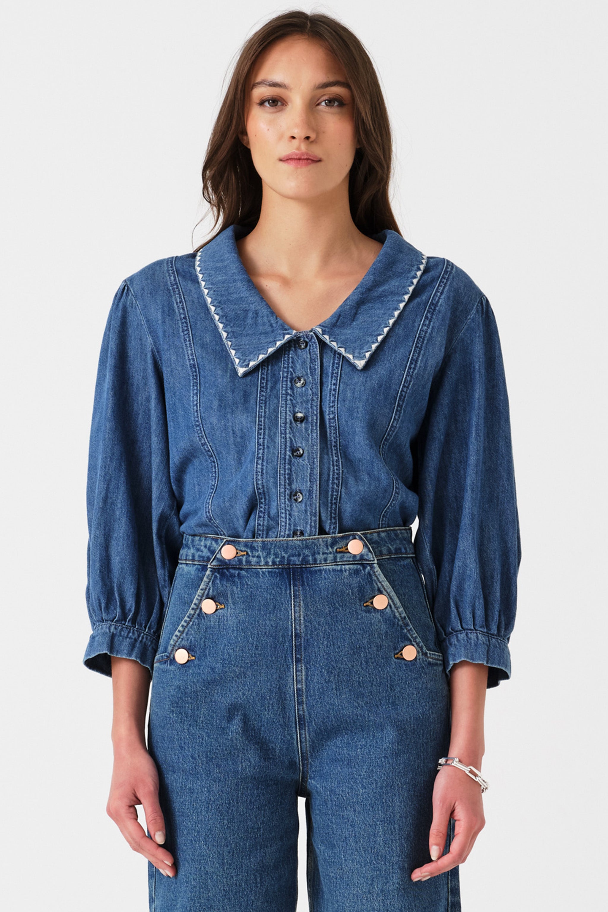 Woman wearing a blue denim button up top with high waisted jeans on a white background.