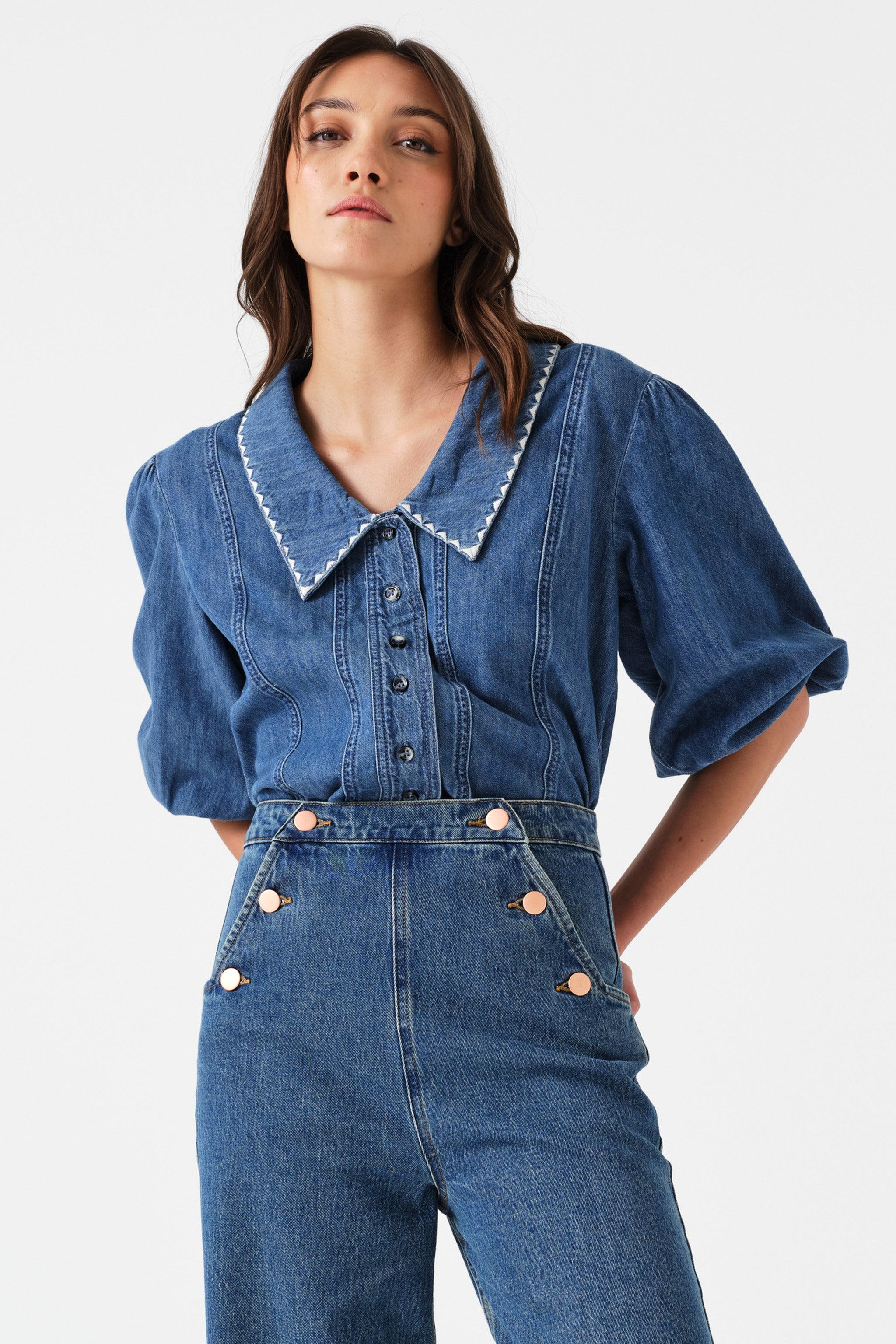 Woman wearing a blue denim button up top with high waisted jeans on a white background.
