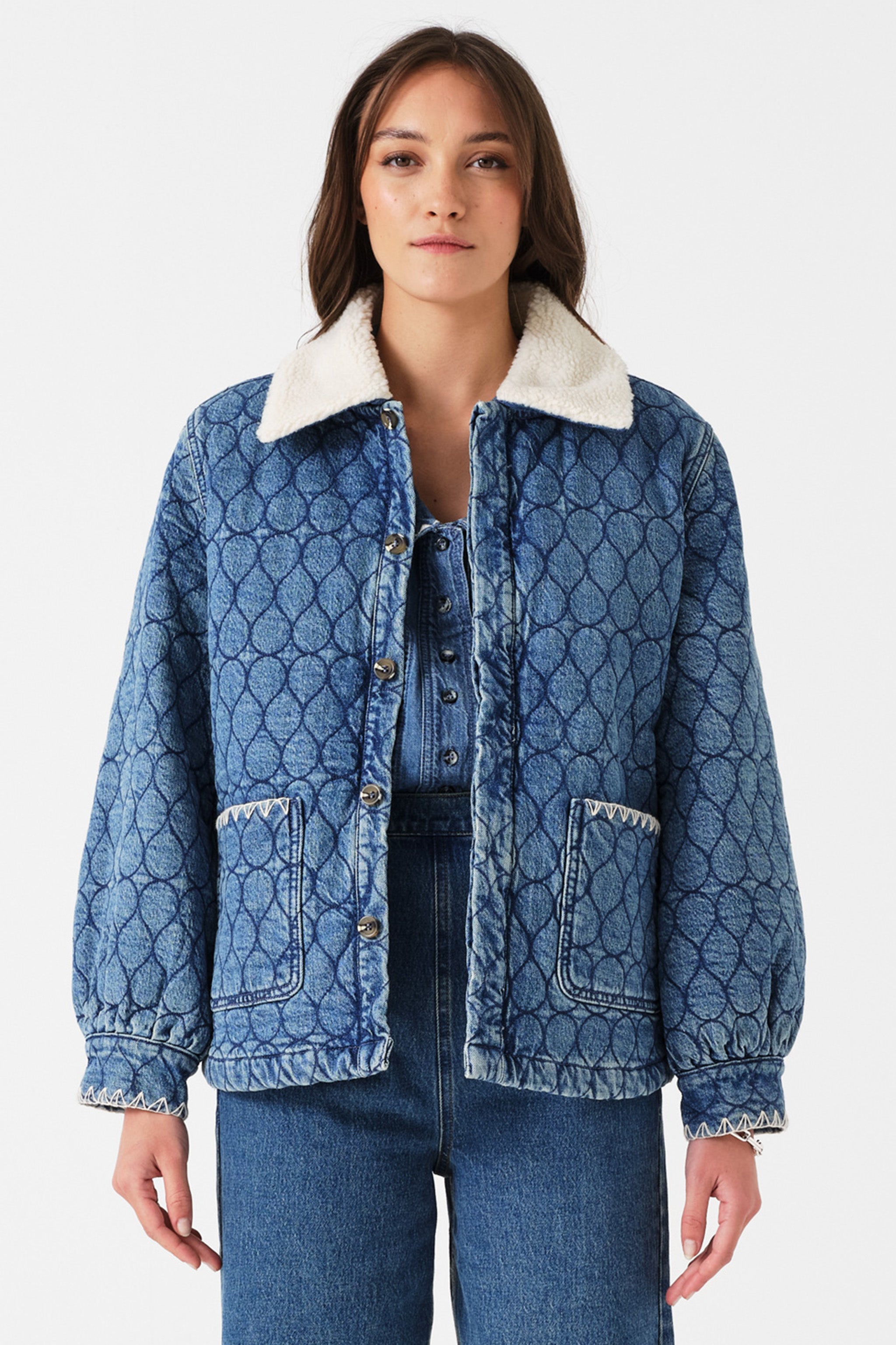 Woman wearing a blue quilted denim jacket with a removable white sherpa collar, blue shirt, and blue jeans on a white background