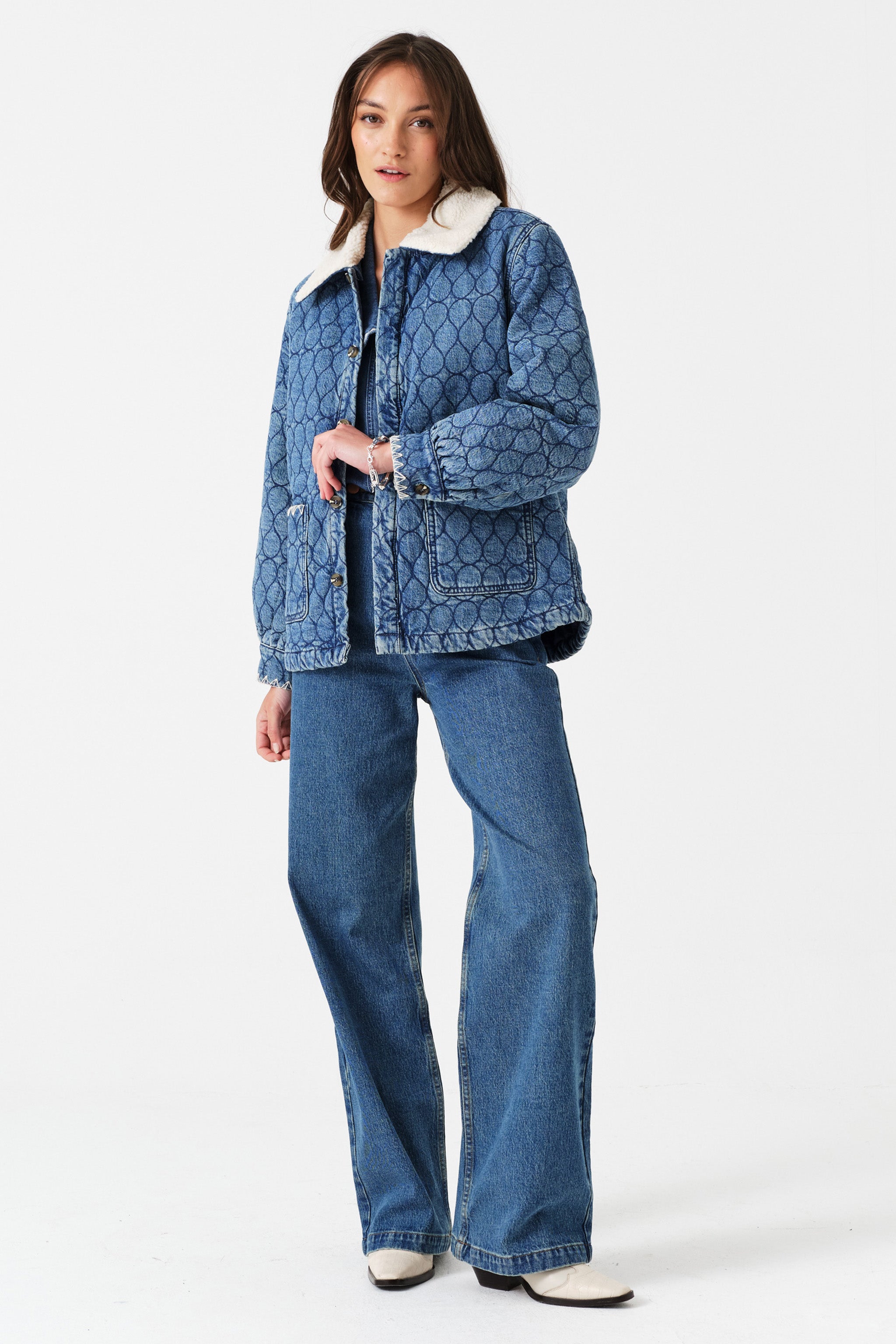 Woman wearing a blue quilted denim jacket with a removable white sherpa collar, blue shirt, and blue jeans on a white background