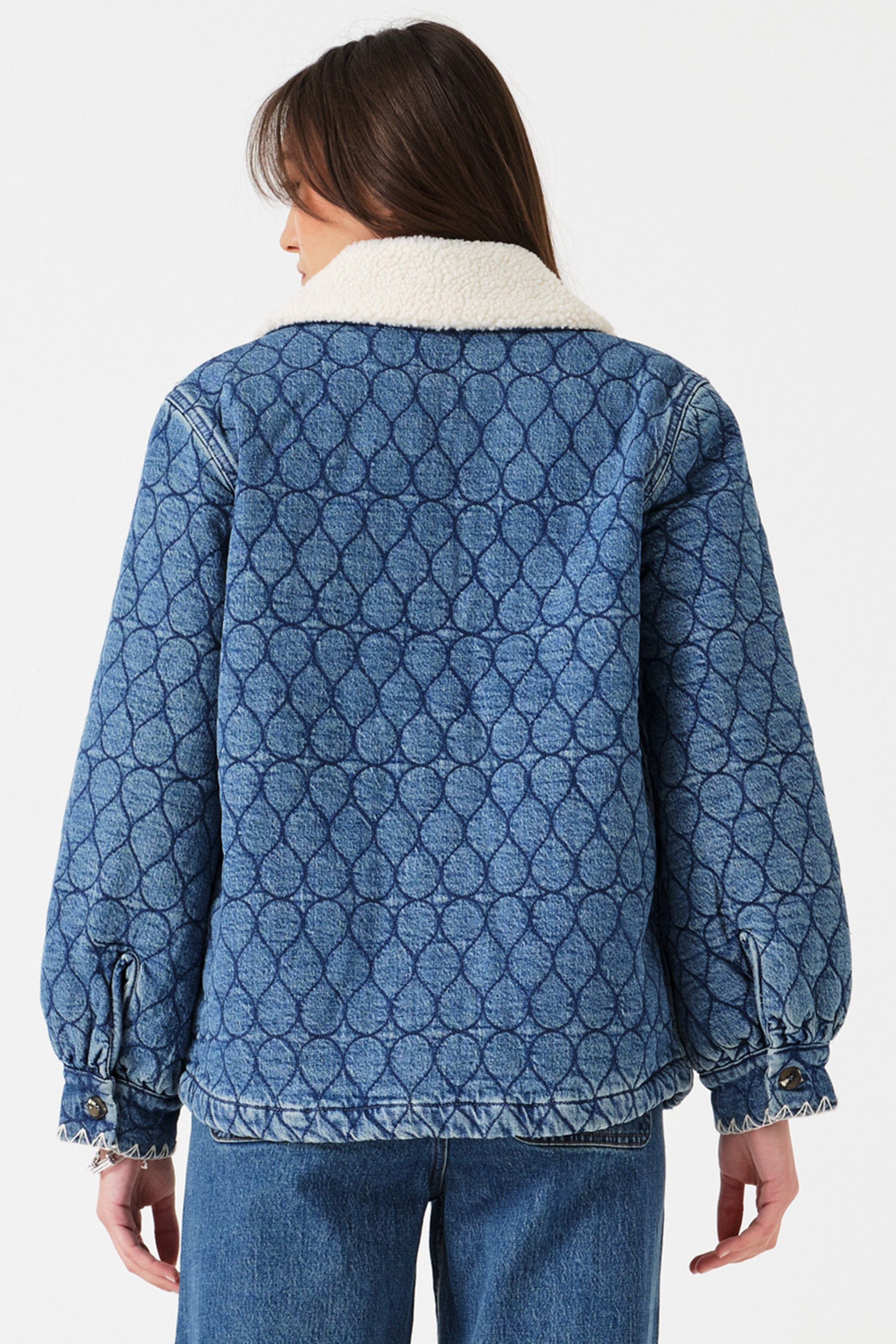 Woman wearing a blue quilted denim jacket with a removable white sherpa collar and blue jeans on a white background