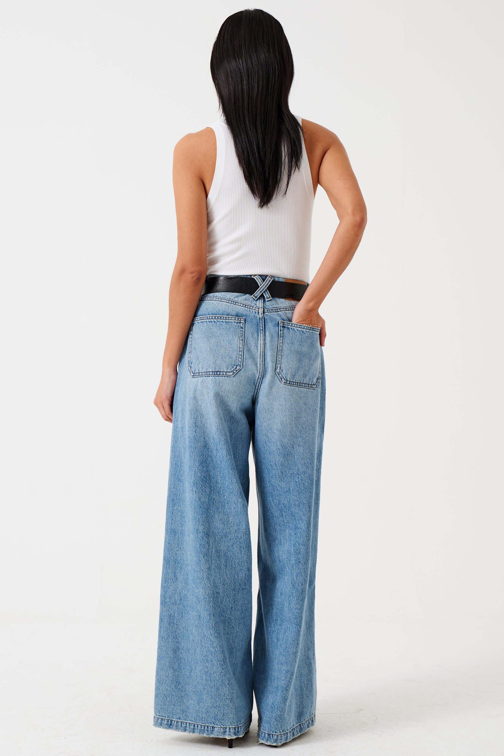 A woman wearing a high-rise, wide-leg, light wash blue denim jean and white shirt with a belt and black shoes against a white background