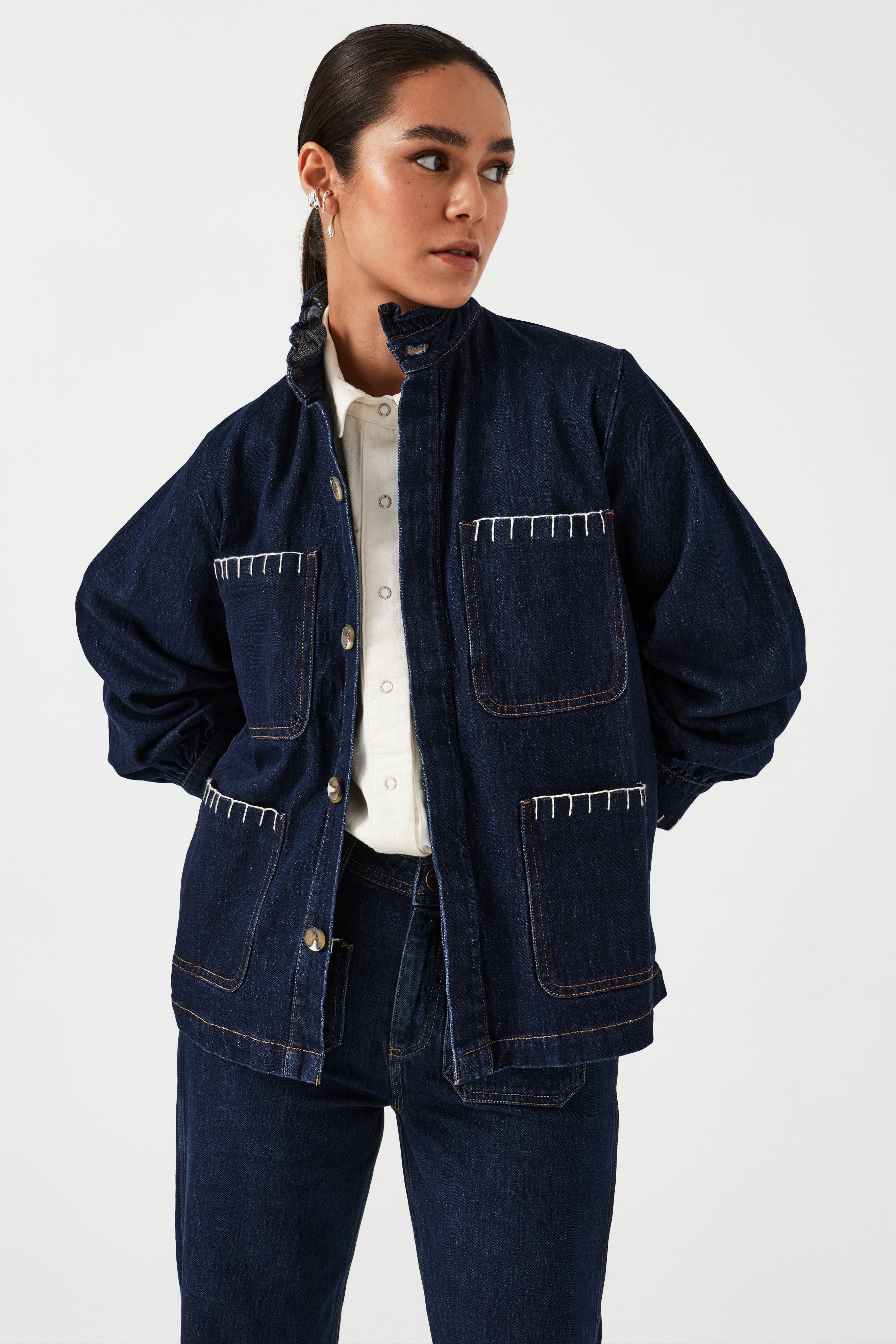 A woman wearing a dark blue vintage style jacket with a frill collar and our signature white blanket stitching on four large patch pockets, paired with a dark blue jeans and a white top against a white background