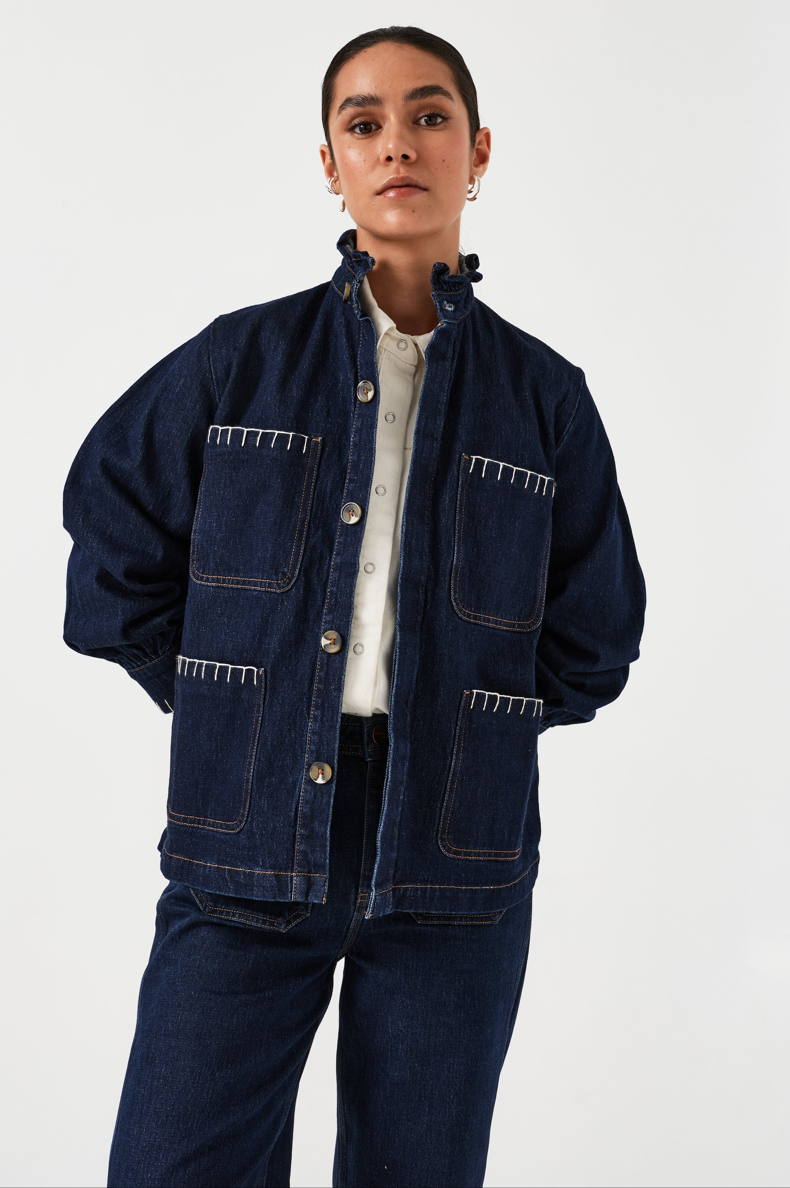 A woman wearing a dark blue vintage style jacket with a frill collar and our signature white blanket stitching on four large patch pockets, paired with a dark blue jeans and a white top against a white background