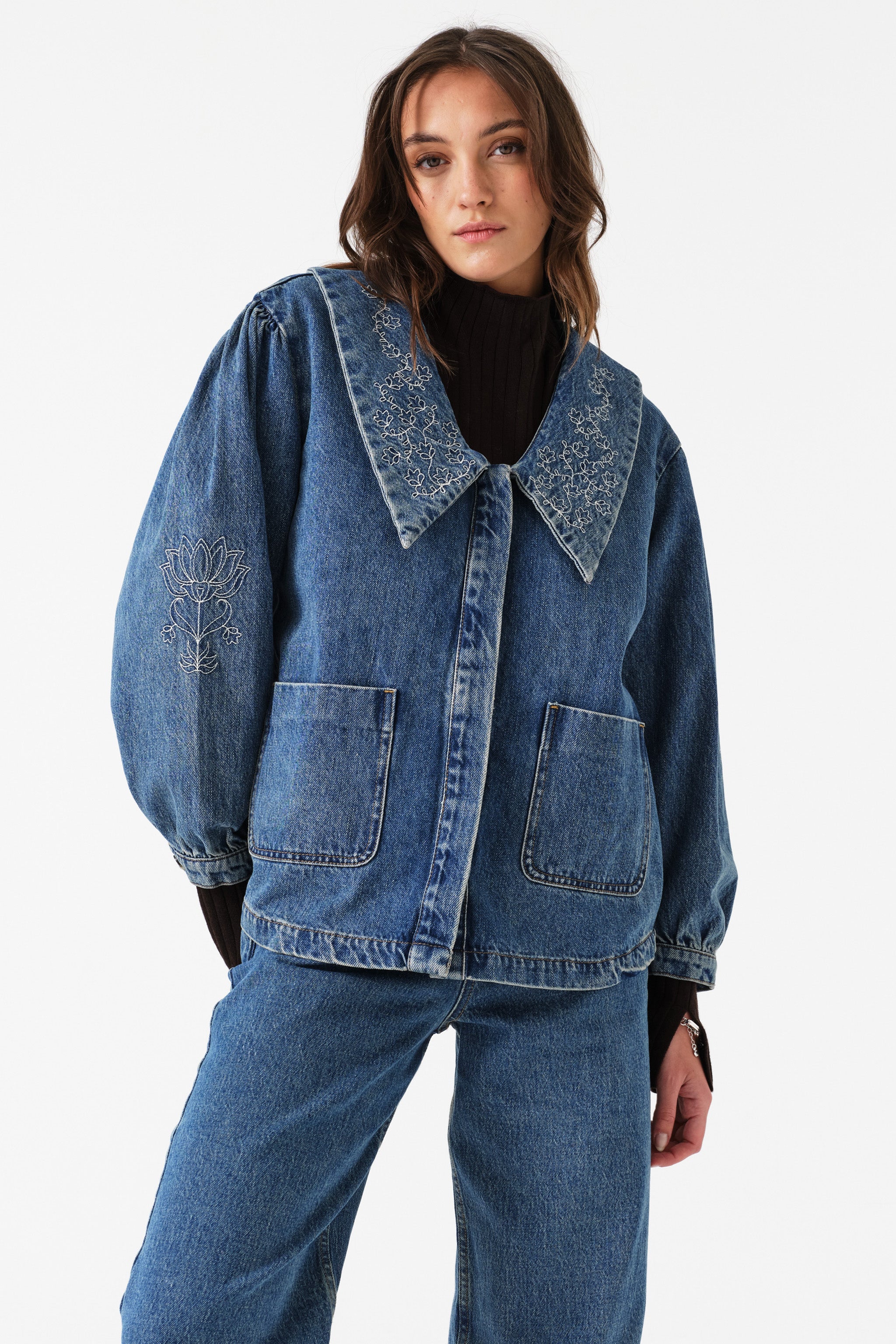 Woman wearing a blue denim jacket with balloon sleeves, embroidery, and pockets with blue jeans on a white background