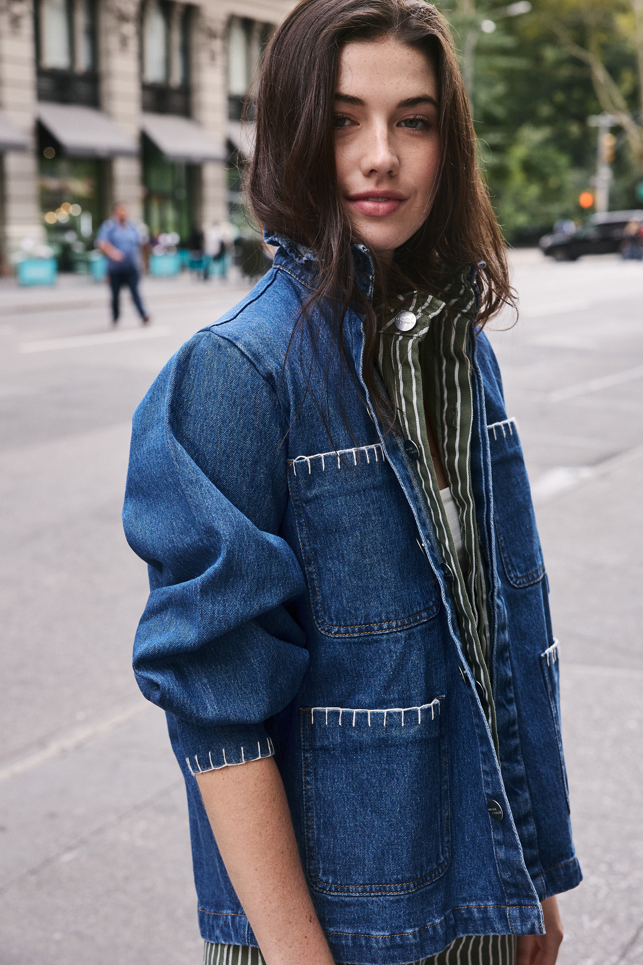 woman wearing a blue denim jacket with a frill collar, white stitch detailing, button-up front, and four large pockets with a green and white striped denim shirt and jeans on a new york city street