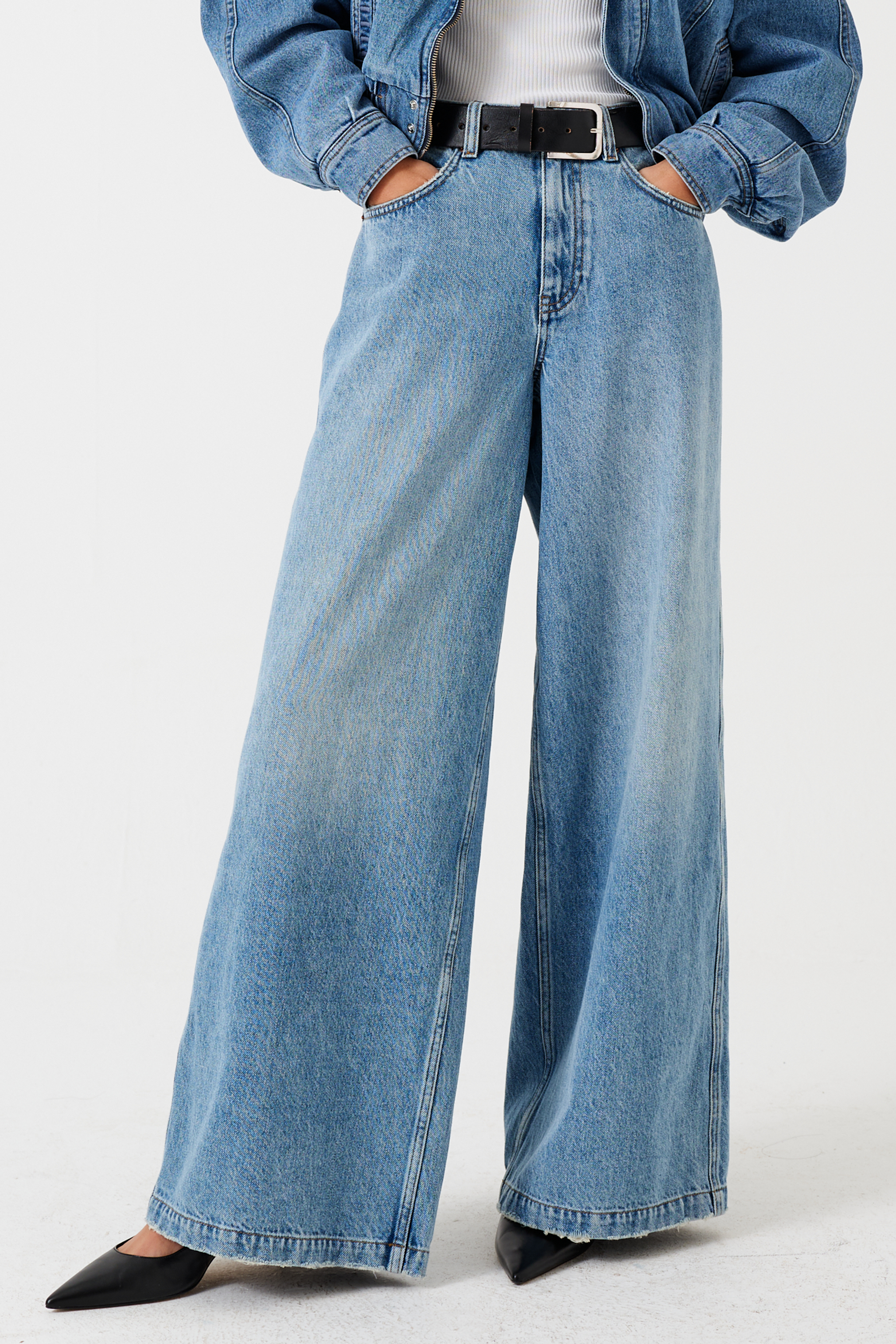 A woman wearing a high-rise, wide-leg, light wash blue denim jean with a belt and black shoes against a white background